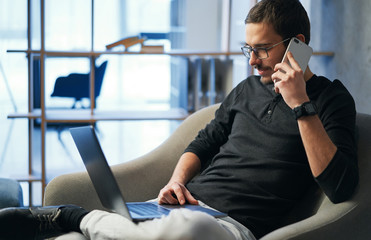 Young man working with phone and computer, receiving phone call, talking with partners 