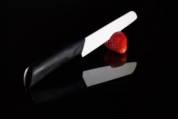A white blade knife cuts a fresh strawberry and its reflection with black background.