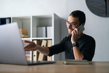 Young man working with phone and computer, receiving phone call, talking with partners 