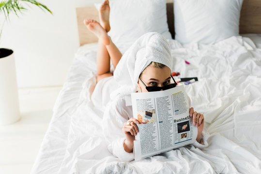 stylish barefoot woman in shirt, sunglasses, jewelry and with towel on head reading business newspaper in bed