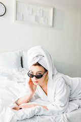 stylish woman in bathrobe and sunglasses, towel and jewelry lying in bed