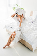 stylish barefoot woman in bathrobe and sunglasses, towel and jewelry sitting on bed