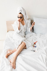 stylish woman in bathrobe and sunglasses, towel and jewelry lying in bed and using smartphone