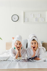 shocked stylish women in bathrobes, earrings and with towels on heads reading magazine while lying in bed