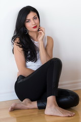 Vertical shot of beautiful sultry long haired woman in sleeveless white top and tight black pants sitting on hardwood floor against plain white background