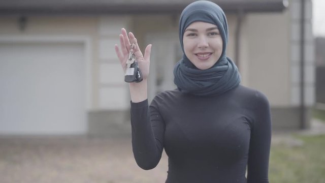 Portrait of independent successful confident smiling young muslim woman holding and waving car keys wearing traditional headscarf outdoors.