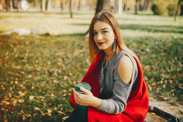 pretty cute girl with make up, dressed in gray sweater sitting in a autumn park on a red plaid and drinking a coffee