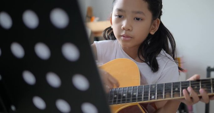 Asian little girl learning to play basic guitar by using acoustic guitar for beginner music instrumental self studying at home