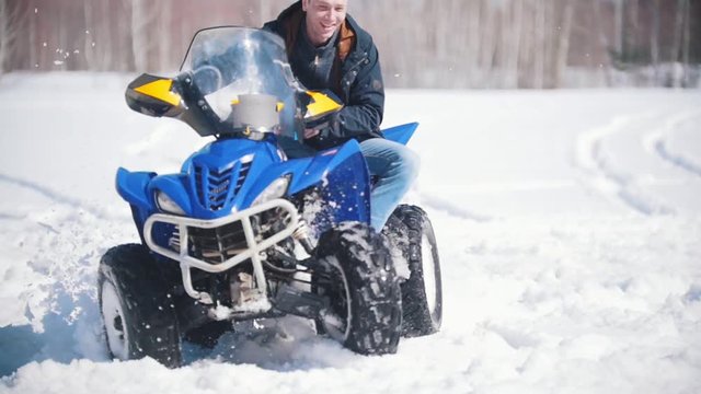 A winter forest in daylight. An adult man riding a big blue snowmobile