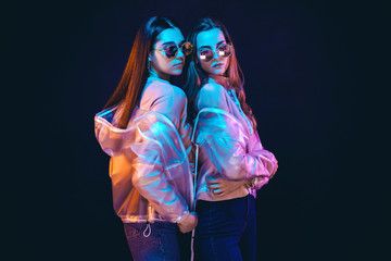 Side view of provocative teen girls in stylish jackets and sunglasses standing on black background in neon illumination
