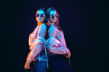 Fototapeta na wymiar Side view of provocative teen girls in stylish jackets and sunglasses standing on black background in neon illumination