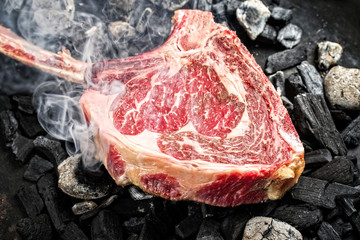 Traditional raw dry aged wagyu tomahawk steak on hot burning charcoal with smoke as closeup on a...