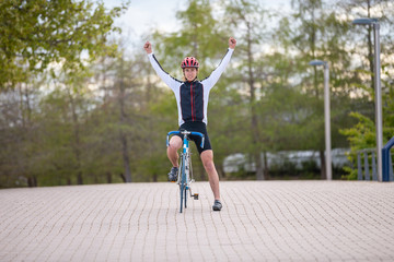 Fototapeta na wymiar Young man in helmet and sportswear raising hands and celebrating victory while riding bicycle on pavement in park