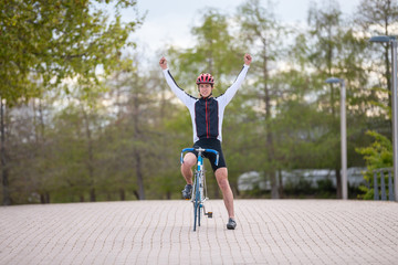 Young man in helmet and sportswear raising hands and celebrating victory while riding bicycle on pavement in park