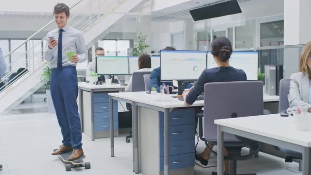 In the Bright Corporate Office Cool Businessman Rides Skateboard through Office Hallway, Uses Smartphone, Shares Stuff on Social Media. In Background Professional People Working on Desktop Computers