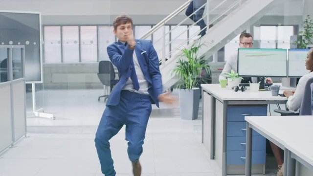 Young Fun Loving and Handsome Businessman Dances Through the Open Space Office Hallway. Worker Celebrating his Latest Achievement. Diverse and Motivated Business People Work on Desktop Computers