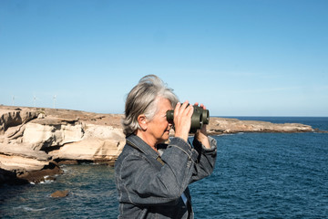 Old woman traveling into Tenerife island looking at the horizon over water with binocular. Exploring and enjoying the nature