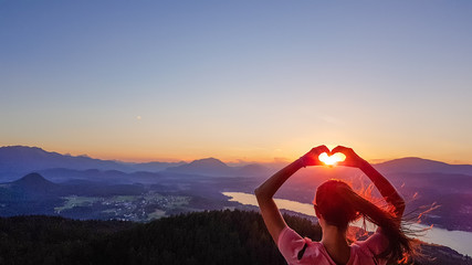 Girl forming a heart with her hands, holding sun in the hands' frame. Below is Wörthersee, Austria.  Soft colors of the sunrise. Share love not hate. Long hair blown by the wind. Pyramidenkogel - obrazy, fototapety, plakaty