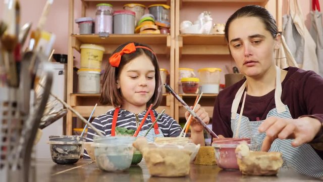 Young girl and teacher at art workshop. Painting clay figures.