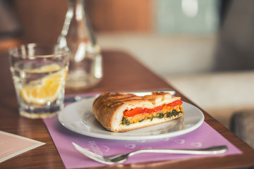 A piece of vegetable pie on a white plate with a fork and a glass of water on the background.