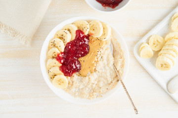 Oatmeal porridge, healthy vegan diet breakfast with strawberry jam, peanut butter, banana, chia on white wooden light background. Top view