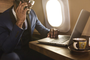 Young attractive and successful African American businessman with glassies talking on the phone and working while sitting in the chair of his private business plane