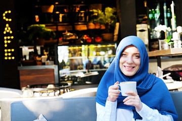 Young beautiful caucasian woman wearing traditional muslim headscarf in hipster coffee shop with big full length windows. Female in blue hijab at cozy cafe. Background, copy space, close up portrait.