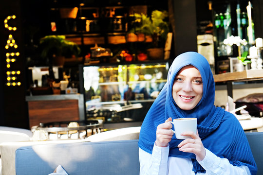 Young beautiful caucasian woman wearing traditional muslim headscarf in hipster coffee shop with big full length windows. Female in blue hijab at cozy cafe. Background, copy space, close up portrait.