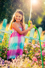Playfull girl watering flowers with rain in the garden at summer day. Child using garden hose on sunny day at sunset lite. Little gardener playing in garden.