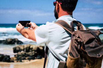 Caucasian man with backpack taking picture with mobile phone technology device to the beach and ocean waves - people enjoying travel and discover hidden place in the world