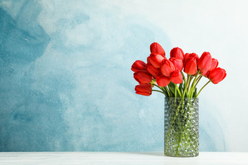 Glass vase with beautiful red tulips on white table against blue background, space for text