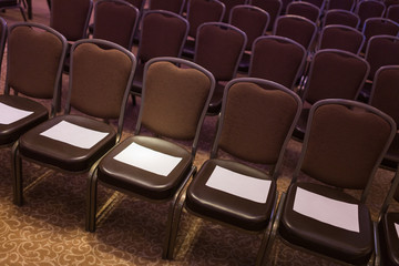 Fototapeta premium Brown chairs the conference hall before the event. First row for special guests. Empty places for your inscriptions on white paper sheets.