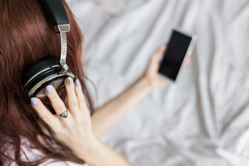 Attractive young brunette woman sits in the morning in her bed with headphones and a smartphone and listens to music in a good mood