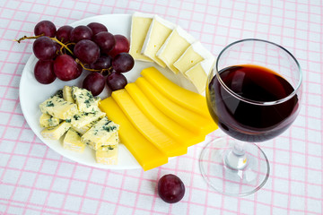 Glass of red wine and white plate with tree kinds of sliced cheese and sweet red grapes. Cheeses covered with edible white and blue mold. Food still life.
