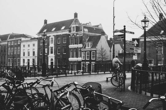 An Elderly Man Riding A Biking And Crossing The Bridge In Harlem, Netherlands. Cyclist Riding Near Bicycle Parking Lot. Vintage Black And White Colors.