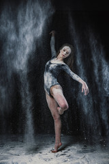 Fototapeta na wymiar A young beautiful girl in a swimsuit and a translucent skirt emotionally dancing on a black background in clouds of white dust