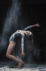 Naklejka na ściany i meble A young beautiful girl in a swimsuit and a translucent skirt emotionally dancing on a black background in clouds of white dust