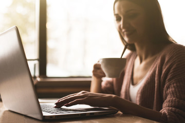 Attractive young woman sits at a table in a cafe with a cup of coffee and enjoys a laptop