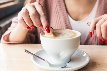 A girl dunks a piece of sugar in coffee. Closeup of hands with red nails make-up and cappuccino cup