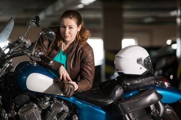 Obraz na płótnie Canvas Biker girl in leather jacket posing on motorcycle in parking lot, blue motorbike and white helmet