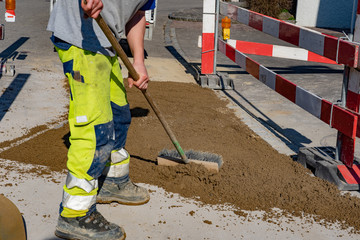 Fototapeta premium Worker with warning trousers and working shoes is scrubbing with a street broom in his hand. Striped boundary close a construction site of a pipeline construction in the street. Close-up. 