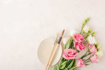 Creative natural composition of blooming pink and white flowers, paint brushes and color palette on beige stone background. Top view, flat lay and copy space. Creative painter workspace