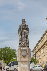 View of the statue of D. Dinis, in the square of the University of Coimbra, surrounding buildings, people and vehicles, cloudy sky, in Coimbra, Portugal