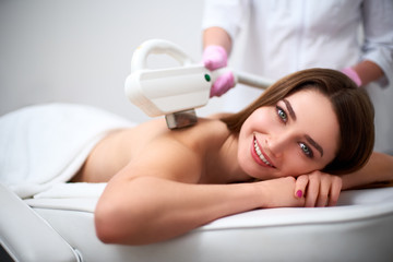 Young pretty smiling woman back laser epilation in beauty salon. Cosmetologist doing depilation...