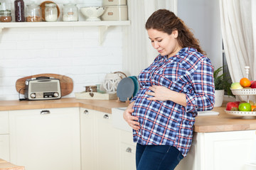 Pretty European pregnant woman holding her belly with hands and looking down at it, kitchen interior