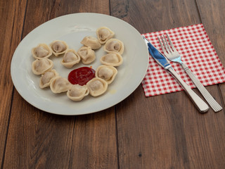 Russian dumplings on a white plate arranged in heart shape and portion of ketchup. Wooden table, Knife and fork,