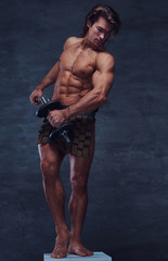 Fototapeta na wymiar Beautiful shirtless bodybuilder is posing with dumbbell on pedestal. He is wearing bandage.
