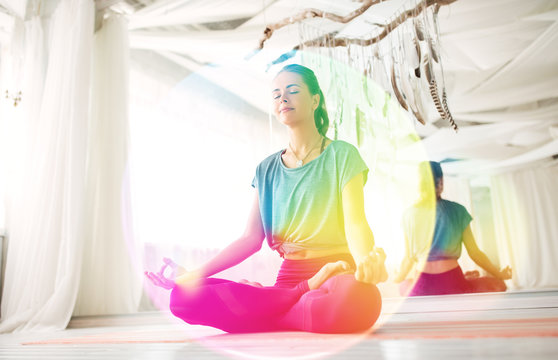 Mindfulness, Spirituality And Healthy Lifestyle Concept - Woman Meditating In Lotus Pose At Yoga Studio Over Rainbow Aura