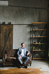 Fototapeta na wymiar Portrait of a man. The groom in a in a gray suit, white shirt and a bow tie is sitting on a brown leather sofa. Room in loft style.