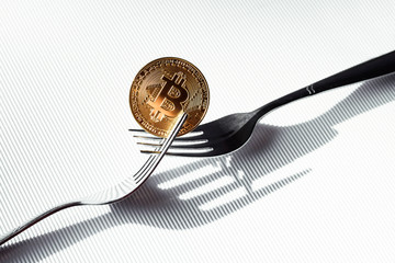 Shiny gold coin with bitcoin symbol between two silver forks on white background. Concept of virtual crypto currency.